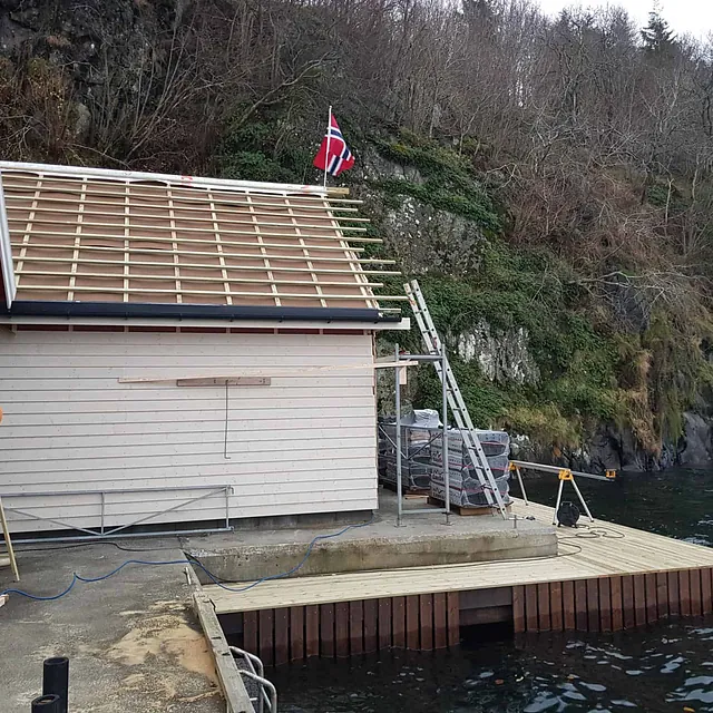 Båthus under bygging ved vannkanten med norsk flagg